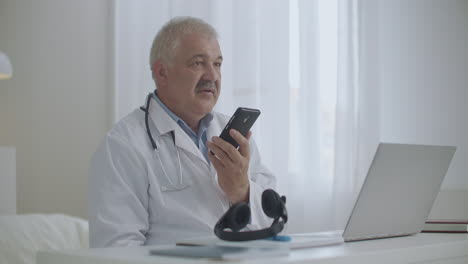 El-Médico-Masculino-Está-Hablando-Por-Altavoz-En-Un-Teléfono-Móvil-En-Su-Oficina-En-La-Clínica-Grabando-Mensajes-De-Voz-Consultando-Al-Paciente-Por-Teléfono-Celular